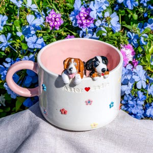 May include: A white ceramic mug with a pink interior and handle, decorated with colourful flowers. Two dog figurines are perched on the rim. The mug has the names "Astor" and "Sadie" with a heart symbol.