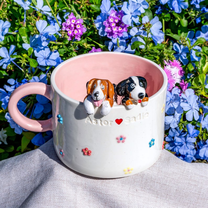 May include: A white ceramic mug with a pink interior and handle, decorated with colourful flowers. Two dog figurines are perched on the rim. The mug has the names "Astor" and "Sadie" with a heart symbol.