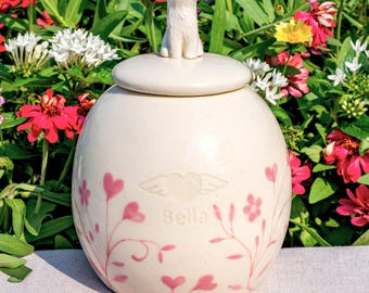 May include: A cream-coloured ceramic urn with a lid topped by a small white dog figurine. The urn features a painted design of pink hearts and floral vines, with the name Bella and a heart with wings engraved on the front centre surface.