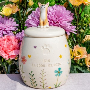 May include: A cream-colored ceramic pet urn with a lid, adorned with a small dog figurine. The urn features a paw print, floral designs, and the name "Jax" with dates. The background is filled with colorful flowers.