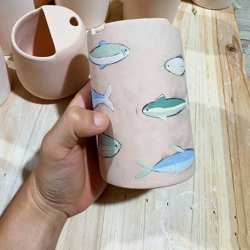May include: A cylindrical ceramic vessel with a matte light pink finish, decorated with hand-painted illustrations of small fish in shades of blue, green, and white. The surface features a smooth texture and a simple, open-top design.