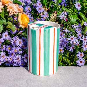 May include: A cylindrical ceramic vase with a white base, featuring vertical stripes of teal and thin red lines. The vase has a rectangular opening at the top. The background includes colorful flowers.