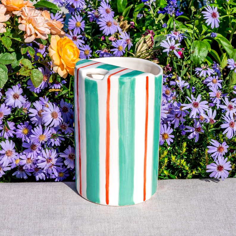 May include: A cylindrical ceramic vase with a white base, featuring vertical stripes of teal and thin red lines. The vase has a rectangular opening at the top. The background includes colorful flowers.