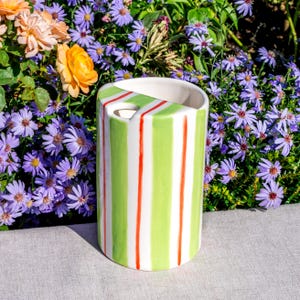 May include: A cylindrical ceramic vase with a white interior, featuring vertical stripes of green, white, and red. The vase has a curved cutout on the top. The background includes purple and orange flowers.