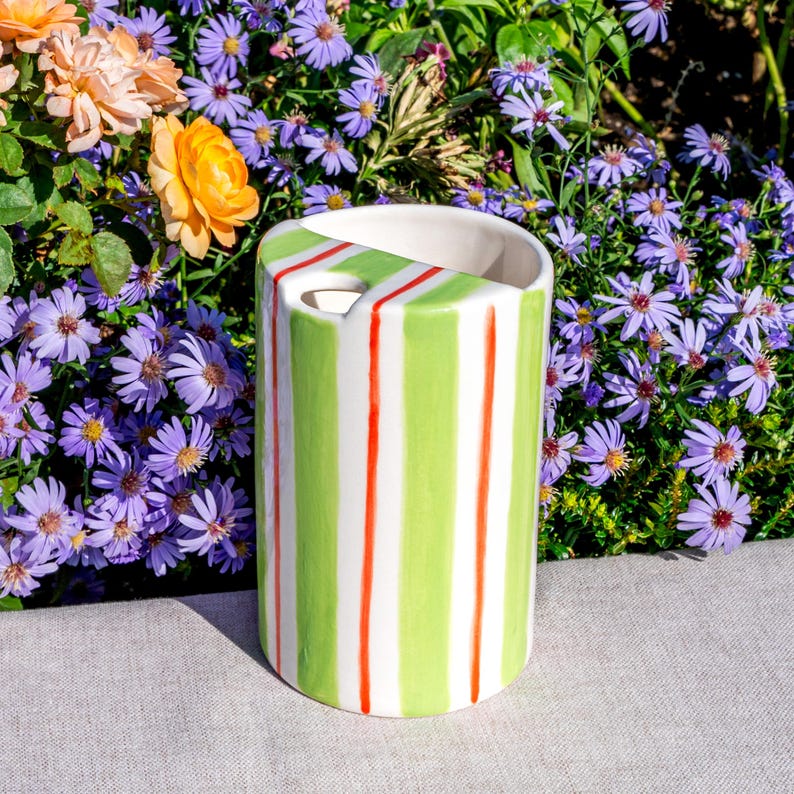 May include: A cylindrical ceramic vase with a white interior, featuring vertical stripes of green, white, and red. The vase has a curved cutout on the top. The background includes purple and orange flowers.