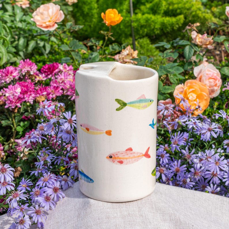May include: A cylindrical off-white ceramic vessel featuring a hand-painted pattern of small, colorful fish in shades of pink, yellow, orange, and blue scattered across the exterior surface, with a smooth finish and a wide, open top opening.