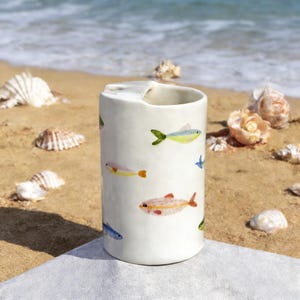 May include: A cylindrical off-white ceramic vessel featuring a hand-painted pattern of small, colorful fish in shades of yellow, orange, pink, and blue. The container has a smooth, slightly irregular surface texture and a wide, open top rim.