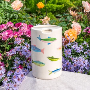 May include: An off-white ceramic vase with a cylindrical shape, decorated with hand-painted fish in various colours. The fish designs include blue, green, yellow, and pink. The vase has a rectangular opening at the top.