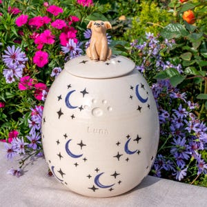 May include: A cream ceramic pet urn with a lid, topped with a small dog figurine. The urn is adorned with blue crescent moons, stars, and a paw print. The name "Luna" is inscribed on the urn.