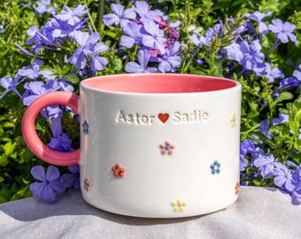 May include: A white ceramic mug with a pink interior and handle, featuring small embossed colorful flowers and the text Astor heart Sadie. The mug has a smooth finish and is decorated with tiny yellow, blue, and orange floral accents.