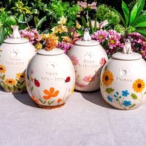 Peut inclure: Quatre urnes funéraires pour animaux de compagnie en céramique crème, chacune surmontée d'une figurine de chien. Les urnes sont décorées de motifs floraux, d'empreintes de pattes et de noms d'animaux avec des dates. Fleurs colorées en arrière-plan.