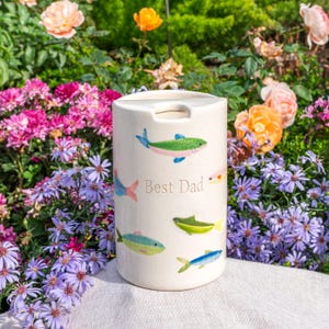 May include: A cylindrical ceramic container with a cream-colored finish, featuring hand-painted illustrations of various small fish in shades of green, blue, and pink. The text "Best Dad" is printed in a simple font on the front surface.