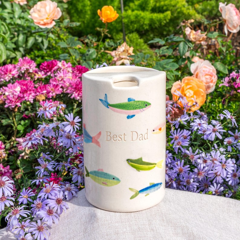 May include: A cylindrical ceramic container with a cream-colored finish, featuring hand-painted illustrations of various small fish in shades of green, blue, and pink. The text "Best Dad" is printed in a simple font on the front surface.