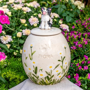 May include: A cream ceramic pet urn with a lid, decorated with hand-painted daisies and the name "FIONA." A small black and white dog figurine sits on the lid. The urn is set against a backdrop of vibrant flowers.