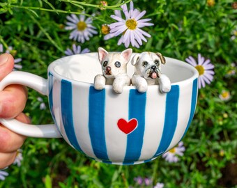 May include: A white teacup with blue vertical stripes and a red heart, held by a hand. Two small dog figurines are perched on the rim. The background features green foliage and white and yellow flowers.