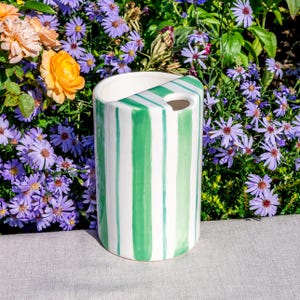 May include: A cylindrical ceramic vase with vertical green stripes on a white background. The vase has a unique angled top with a small hole. The vase is set against a backdrop of purple and orange flowers.