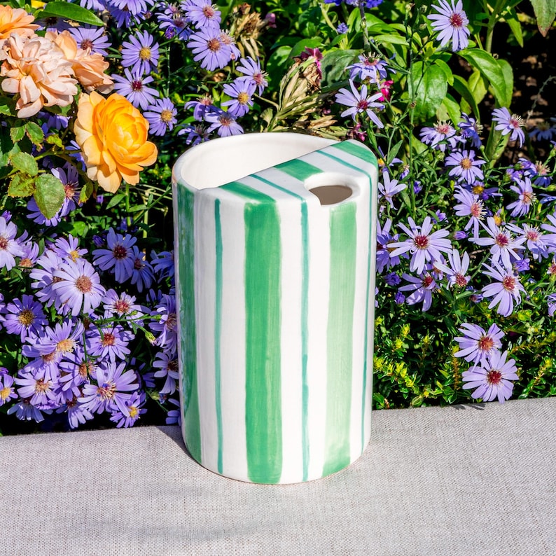May include: A cylindrical ceramic vase with vertical green stripes on a white background. The vase has a unique angled top with a small hole. The vase is set against a backdrop of purple and orange flowers.