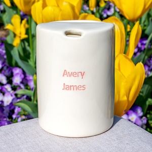 May include: A cream ceramic candle holder with the name "Avery James" in pink lettering. The cylindrical candle holder has a small opening at the top. The background features yellow tulips and purple flowers.