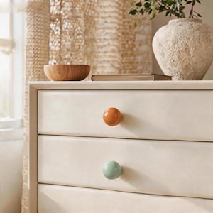 May include: A light cream-colored dresser featuring two drawers, each equipped with a single round knob. The top drawer knob is a glossy burnt orange, while the bottom drawer knob is a smooth, matte sage green finish on the drawer fronts.