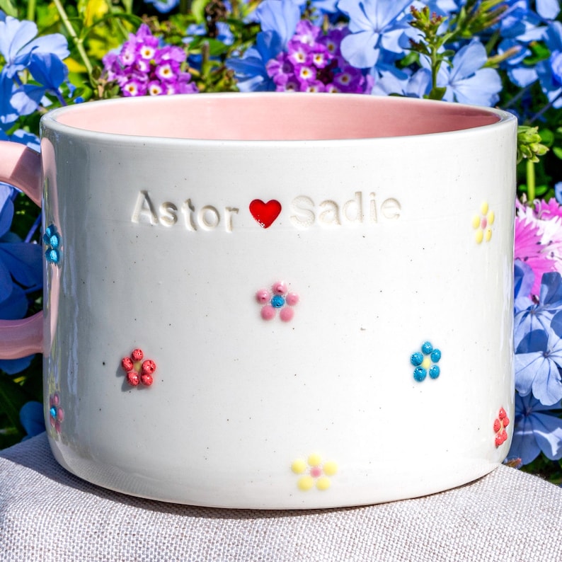 May include: White ceramic mug with a pink interior and handle, decorated with colourful floral details. The mug has the names "Astor" and "Sadie" separated by a red heart. A charming and personalised gift.
