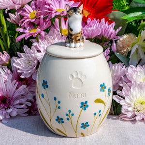 May include: A cream ceramic pet urn with a lid, decorated with a paw print and the name "Nunu." It has blue floral patterns and is topped with a small cat figurine. The background is filled with colourful flowers.