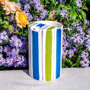 May include: A cylindrical ceramic vase with vertical stripes in blue, green, and purple. The vase has a rectangular opening at the top. The background features vibrant purple flowers and greenery.