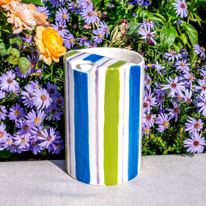 May include: A cylindrical ceramic vase with vertical stripes in blue, green, and purple. The vase has a rectangular opening at the top. The background features vibrant purple flowers and greenery.