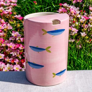 May include: A cylindrical ceramic utensil holder with a glossy light pink finish, decorated with hand-painted blue fish featuring yellow fins. The top edge has a semi-circular cutout, and the interior is white, providing a smooth, clean surface.