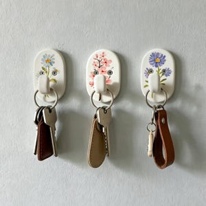 May include: Three oval wall hooks with white ceramic bases, each featuring a unique floral design. The left hook displays blue daisies, the centre hook shows pink cherry blossoms, and the right hook depicts purple asters. Each hook holds a set of keys.