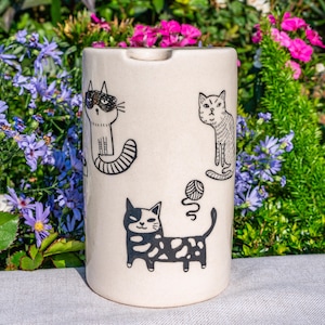 May include: A cream ceramic ashtray with black cat illustrations. The cylindrical ashtray has a small notch. The cats are depicted in different poses, including one with a ball of yarn. The background is blurred with flowers and foliage.