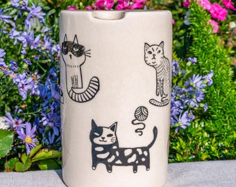 May include: A cream-colored ceramic ashtray featuring black line drawings of cats. The ashtray has a cylindrical shape with a small notch. The cats are in various poses, with one playing with a ball of yarn. The background is blurred with flowers.
