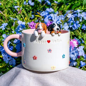 May include: White ceramic mug with a pink handle and interior, decorated with colourful floral accents. Two dog figurines sit on the rim. The mug is inscribed with "Astor ❤️ Sadie."