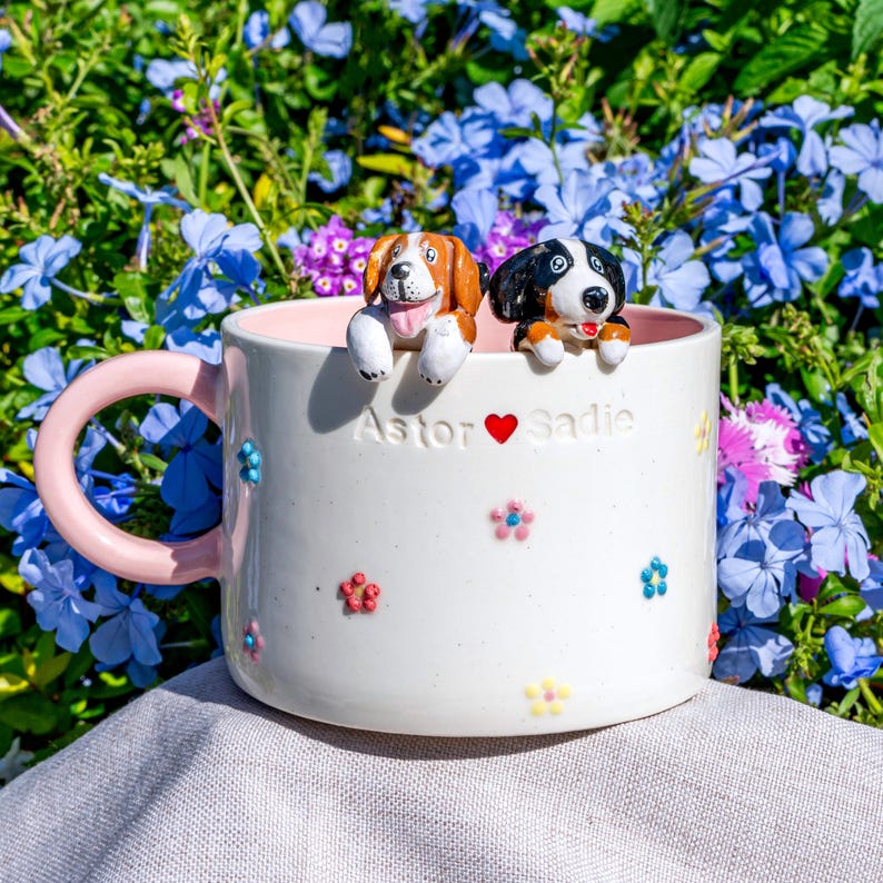 May include: White ceramic mug with a pink handle and interior, decorated with colourful floral accents. Two dog figurines sit on the rim. The mug is inscribed with "Astor ❤️ Sadie."