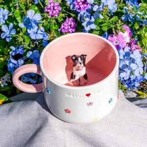 May include: A white ceramic mug with a pink interior and handle, featuring a small dog figurine inside. The mug is decorated with colourful floral accents and the name "Astor" with a heart symbol.