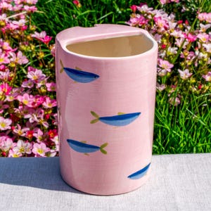 May include: A cylindrical ceramic utensil holder with a glossy light pink finish, featuring a hand-painted pattern of small blue fish with yellow fins scattered across the exterior surface and a wide, open top with a notched rim detail.