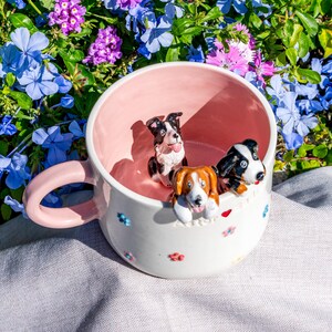 May include: A white ceramic mug with a pink interior and handle, decorated with small, colourful flower designs. Three miniature dog figurines are perched on the rim. The background features vibrant blue and purple flowers.