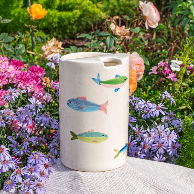 May include: A cylindrical off-white ceramic utensil holder featuring a hand-painted pattern of small, colorful fish in shades of blue, green, and pink. The top edge has a distinct notched cutout, and the surface has a smooth, speckled finish.
