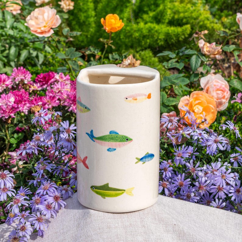 May include: A cylindrical off-white ceramic utensil holder featuring a hand-painted pattern of small, colorful fish in shades of green, blue, pink, and orange scattered across the exterior surface with a smooth, matte finish.