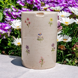 May include: A cylindrical ceramic container with a speckled, off-white finish. The container is adorned with hand-painted, colorful floral designs. The background features a garden with white daisies and purple flowers.