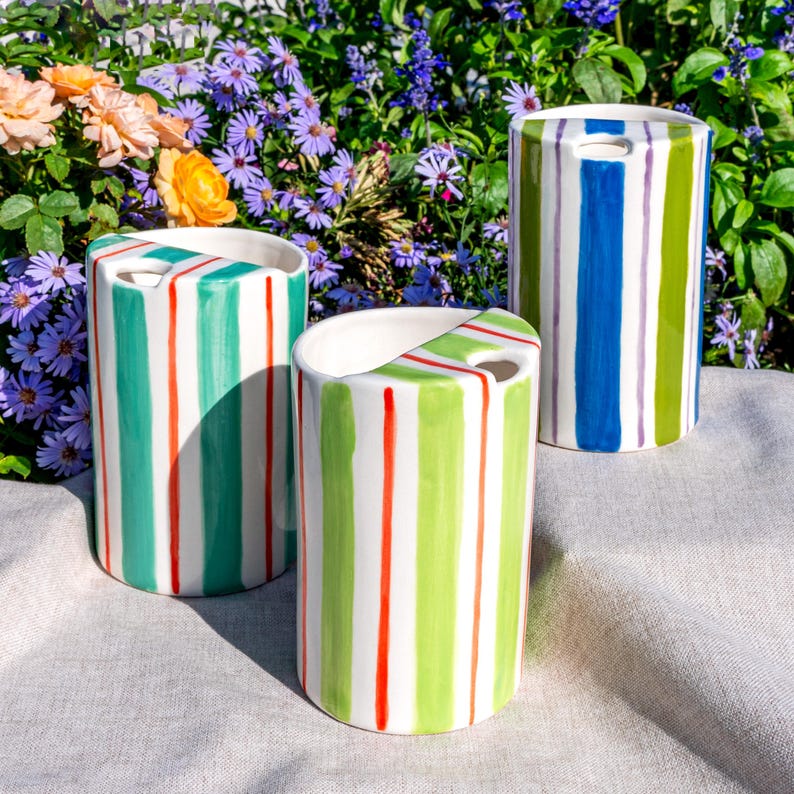 May include: Three ceramic vases with vertical stripes in various colors. The vases have a cylindrical shape with a handle-like opening at the top. The stripes are in shades of green, red, blue, and purple, creating a vibrant and decorative look.