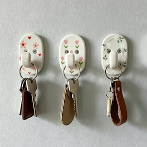 May include: Three oval-shaped wall hooks made of white ceramic, each featuring a unique floral pattern. The left hook has red hearts and pink flowers, the centre has pink tulips, and the right has white daisies. Each hook holds a metal key ring.