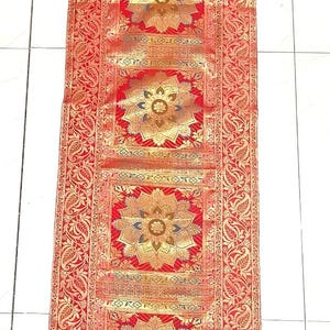 May include: Red and gold decorative table runner with a floral pattern. The runner features a repeating design of gold floral medallions on a red background, with ornate gold borders. The runner is rectangular and appears to be made of a woven fabric.