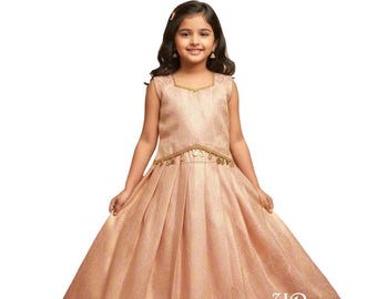 Puede incluir: Una niña pequeña con un vestido sin mangas color melocotón con una falda plisada. El vestido tiene adornos dorados alrededor del cuello y la cintura. La niña sonríe y lleva sandalias marrones.
