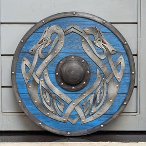 Könnte beinhalten: Ein runder, blauer Holzschild mit silberner Umrandung und Nieten. Der Schild hat einen zentralen Knauf und ein symmetrisches Design aus zwei stilisierten Tierfiguren in Silber mit roten Akzenten. Durchmesser ca. 61 cm.