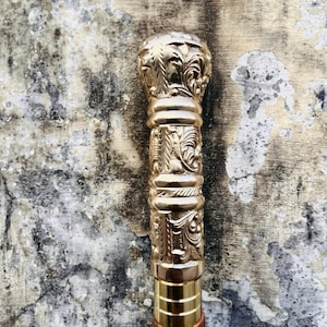 Könnte beinhalten: Goldfarbener Spazierstock mit aufwendigen floralen und Rankenmustern. Der obere Teil des Stocks hat einen abgerundeten, verzierten Griff. Der Schaft besteht aus goldfarbenem Metall und dunkelbraunem Holz. Dekoratives Accessoire.