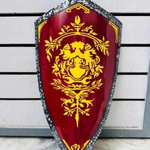 May include: A burgundy shield with a yellow emblem. The shield has a decorative border and a central design featuring a stylized floral and animal motif. The shield is likely made of metal.