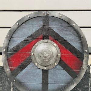 May include: A circular wooden shield with a distressed finish. The shield has a blue and black background with a red stripe design. A metal rim and central boss are secured with rivets, creating a rustic, historical aesthetic.