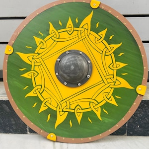Puede incluir: Un escudo redondo de madera con fondo verde y diseño de sol amarillo. El escudo tiene un borde marrón y un centro metálico. Se adjuntan correas de cuero amarillas.