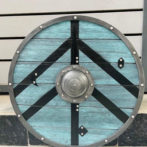 Puede incluir: Un escudo vikingo redondo con una superficie de madera azul claro y un diseño geométrico negro. El escudo tiene un borde de metal y un jefe central de metal. Remaches decorativos y detalles metálicos.