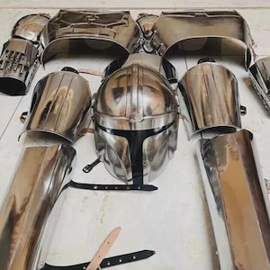 Könnte beinhalten: Eine Sammlung glänzender, silberfarbener Rüstungsteile, darunter ein Helm, eine Brustplatte und Armschützer. Die Rüstung ist auf einer hellen Oberfläche angeordnet, was ihre reflektierende Qualität und ihr aufwendiges Design zeigt. Schwarze Riemen und Schnallen sind ebenfalls sichtbar.